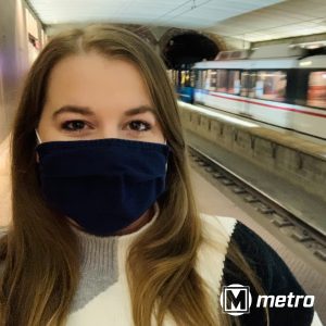 Selfie of a woman wearing a mask on the 8th and Pine MetroLink platform with a train driving past in the background. Metro logo on the bottom right