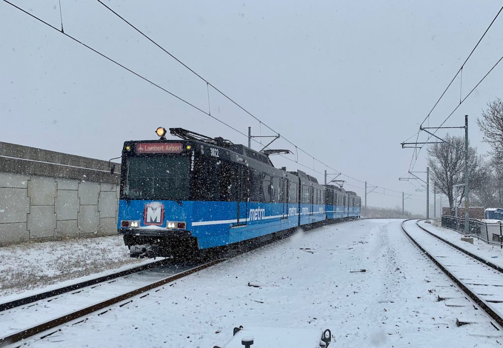 Front of a west-bound MetroLink train arriving at the North Hanley station. It is snowing and snow covers the ground and tracks.