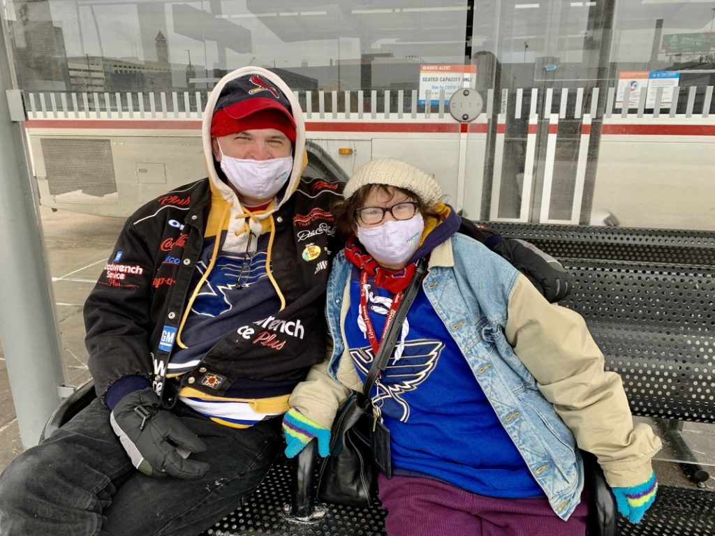 Passengers Tom and Carol sitting at the #10 bus stop at the Civic Center Transit Center. They are both sitting on the bench, smiling at the camera and wearing face masks