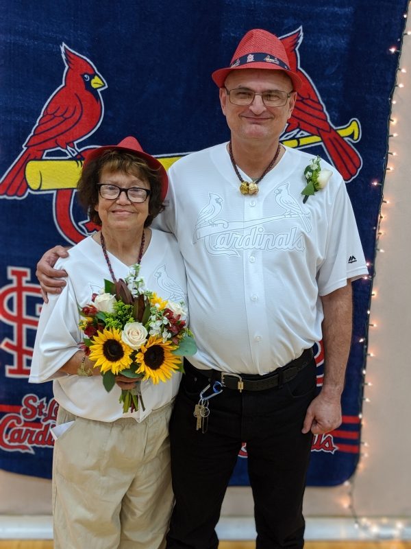 Photo of passenger Tom and Carol on their wedding day, smiling at the camera. Tom's arm is around Carol, and they are wearing matching white Cardinals jerseys. Carol is holding a yellow and purple bridal bouquet. 