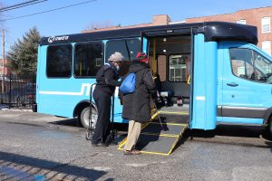 Call-A-Ride operator, Barbara, helps rider, Donna, on board a Call-A-Ride van. The doors are open and the ramp is lowered. Red brick buildings appear in the background.