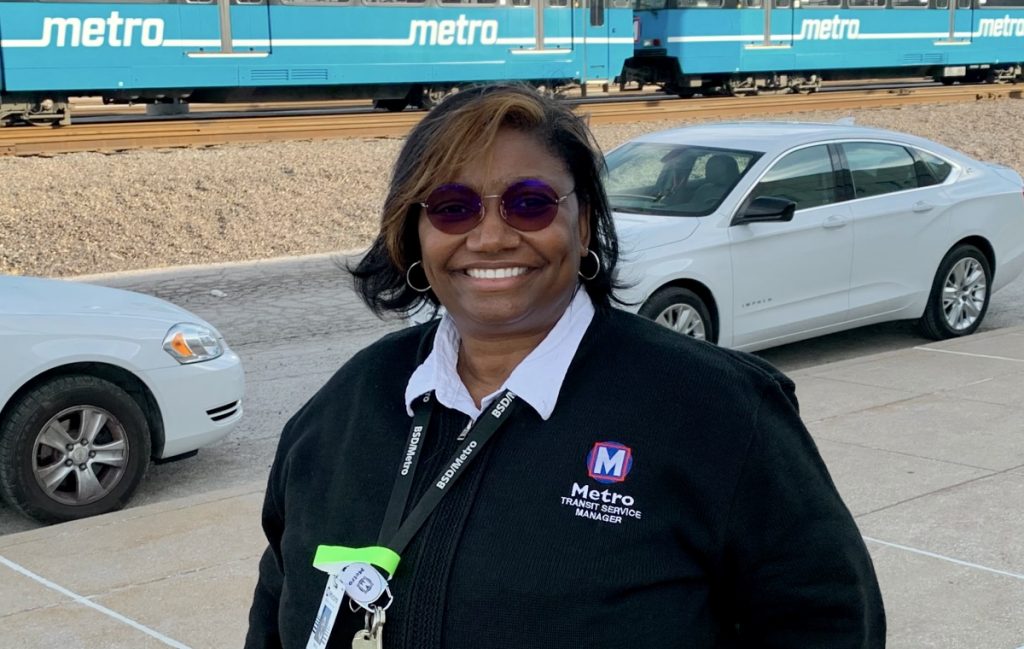 Team member Edith standing outside of a Metro facility, smiling at the camera and wearing glasses. There are two white cars behind her, and in the background is a blue MetroLink train