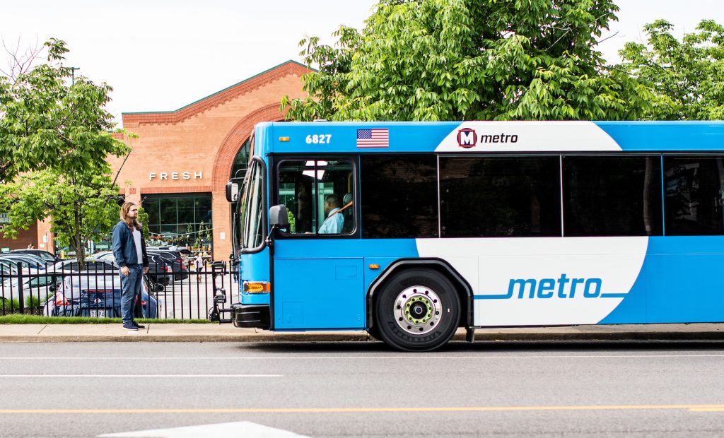 A MetroBus pulls up to a bus stop in front of the Clayton Rd. Schnucks. A rider waits at the bus stop.