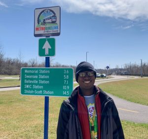 Metro Passenger Dominike standing in front of the MetroBikeLink trail sign at the Fairview Heights MetroLink Station
