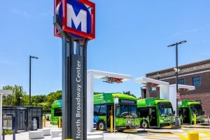 3/4 shot of three electric Metrobuses parked at three charging station in the North Broadway Transit Center. A North Broadway lollipop Metro sign appears in the foreground. The overhead charging pantographs are folded up.