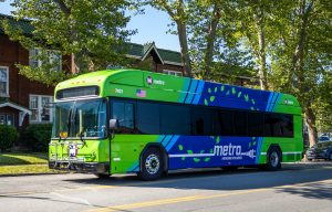 A 40-foot electric bus stops along a residential street. Trees, cars, and housing appear in the background.