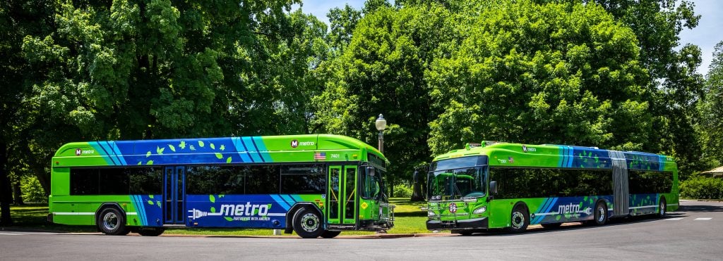 An articulated electric bus and an electric bus face each other in a roundabout in Tower Grove Park. Trees appear in the background.