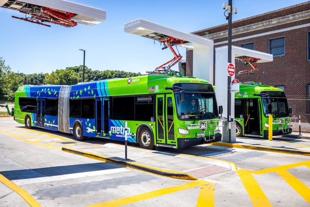 Two articulated electric MetroBuses park at the charging stations at the North Broadway Transit Center. The overhead charging pantographs lower to make contact with the buses.