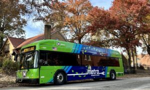 Photo of electric bus on street with trees with orange/red fall leaves in background