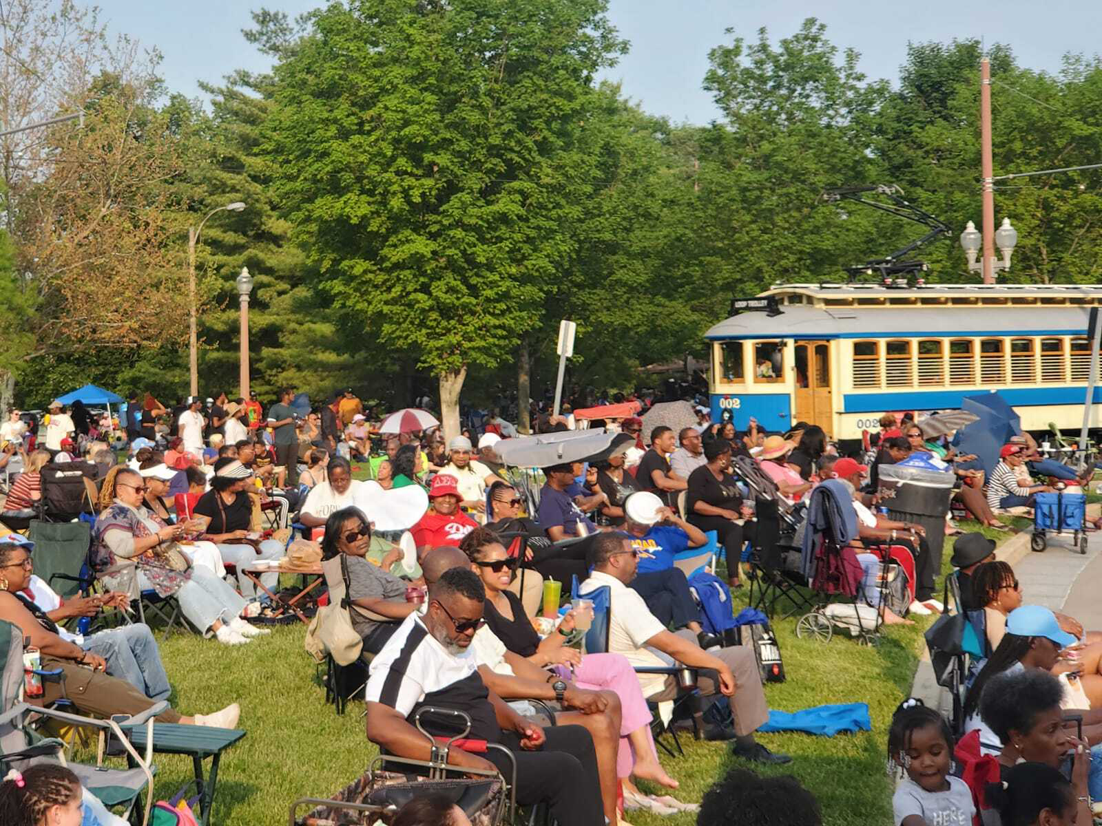 A large group of people seated in chairs and on blankets outdoors in the sun, with a Loop Trolley car and trees in the background