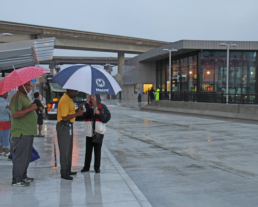 Photo of three individuals standing underneath umbrellas during a rainy day at the Civic Center Transit Center, with the Transit Center building in the background